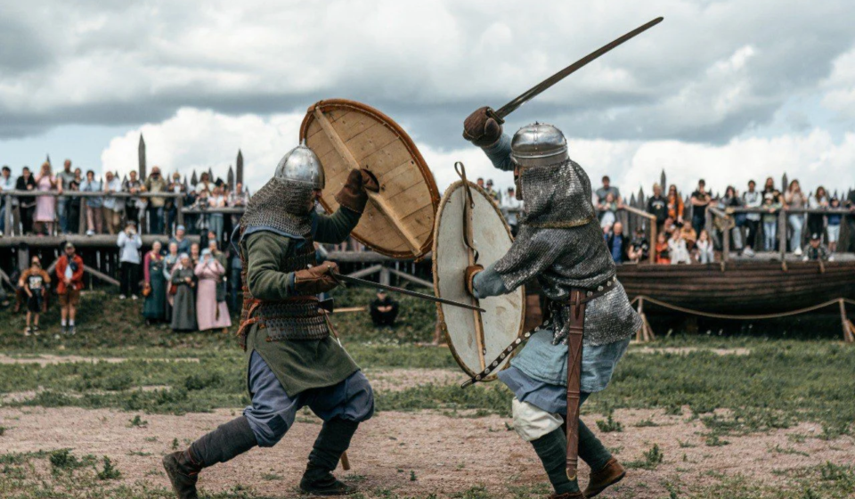 Два воина в исторических доспехах эпохи викингов сражаются на мечах со щитами на арене фестиваля «Kirjálaland» — Карельская земля. На заднем плане — толпа зрителей и ладья.