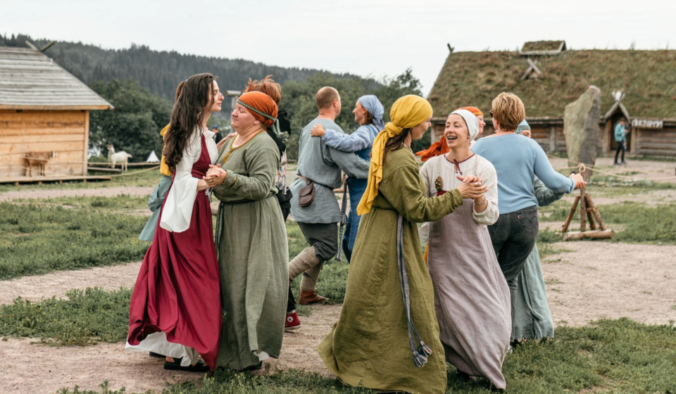Группа людей в исторических средневековых костюмах радостно танцует на фоне деревянных построек и зелёного луга на фестивале «Kirjálaland» — Карельская земля. Уличное театральное представление и живая история.